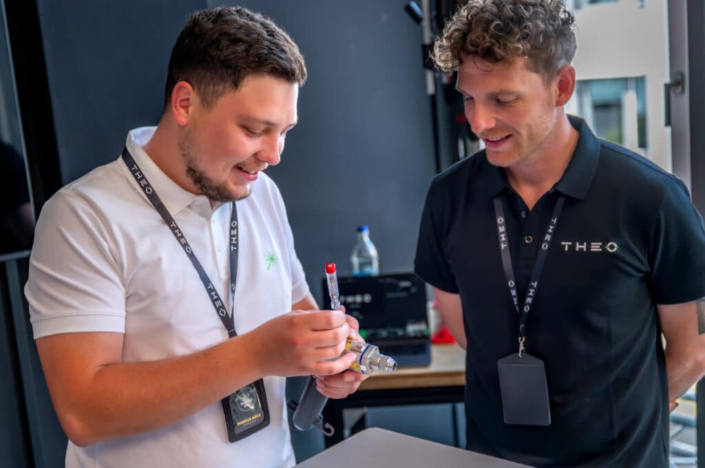 two men standing next to each other, one wearing a white polo and the other a black polo. They both look at a welding torch one of them holds in his hand